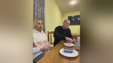 Media: A video shows a young blonde woman in a pink-striped hoodie, and a bald man in a black hoodie, eating at a wooden table with a sandwich and a blue cup. The background has yellow walls and a TV.