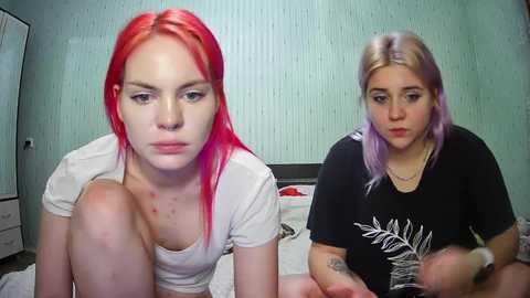 Media: Video of two young women with pink and purple hair, seated on a bed in a room with light blue walls. One wears a white top, while the other wears a black shirt.