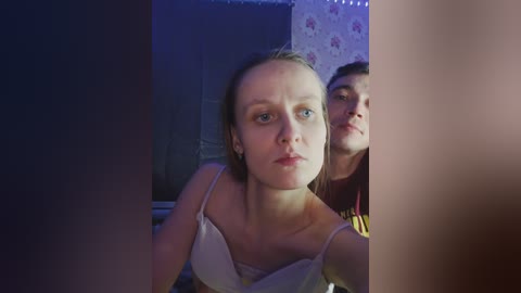 Video of a blonde woman with light skin in a white bra, looking directly at the camera, with a dark-haired man behind her, in a dimly lit room with floral wallpaper.