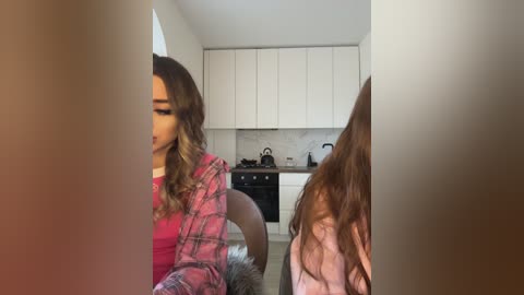 Media: Video of a woman with long, wavy brown hair, wearing a pink plaid shirt, sitting on a chair in a modern kitchen. A woman with long, straight brown hair is seen from behind.