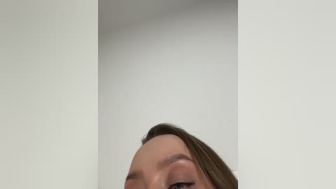 Video of a partially visible woman with light brown hair and fair skin, captured from an upward angle, emphasizing her nose and upper lip, against a plain white wall.
