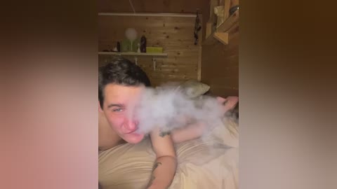 Video of a shirtless, fair-skinned, tattooed man with short dark hair, lying face down on a bed, exhaling a visible cloud of smoke. Background features a wooden-paneled room with a shelf holding personal items.