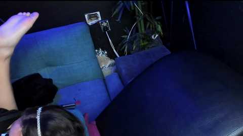A video of a person in a dimly lit room, lying on a blue couch with a black cushion. The person's feet are up, wearing white socks. There's a plant and a phone charger nearby.