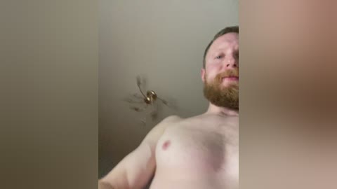 A video of a shirtless, fair-skinned man with a beard and brown hair, standing against a beige wall. A cloud of dust or smoke obscures part of his face and upper body, creating a sense of movement and mystery.