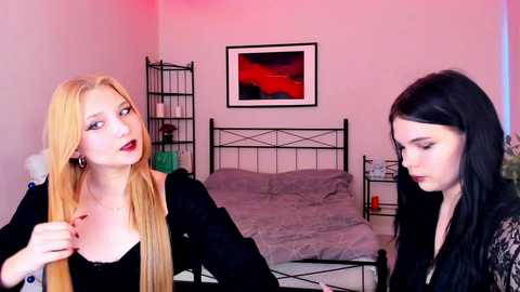 Video of two young women in a bedroom; one blonde with fair skin, black dress, and red lipstick; the other pale-skinned with black hair, black lace top, and dark lipstick. Background includes a black metal bed, red wall, and abstract artwork.
