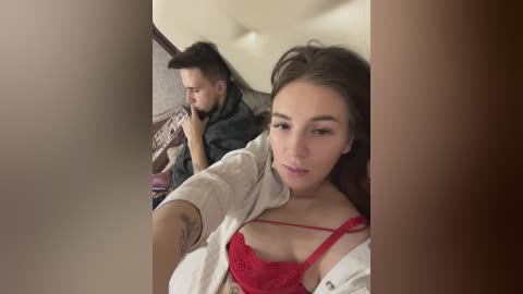Media: Video of a young woman with fair skin, brown hair, and a red lace bra, taking a selfie in a dimly lit room with a man in the background.