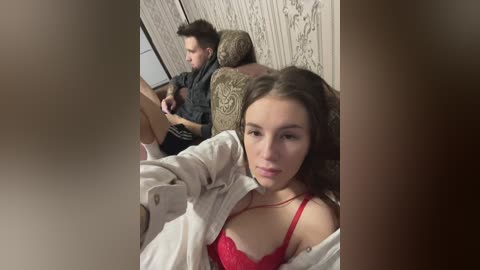 Media: A video captures a young woman with long brown hair, wearing a red lace bra, sitting on a beige sofa. She has a neutral expression. Behind her, a young man in a black shirt and shorts is seated, looking at his phone. The background features a floral wallpaper.