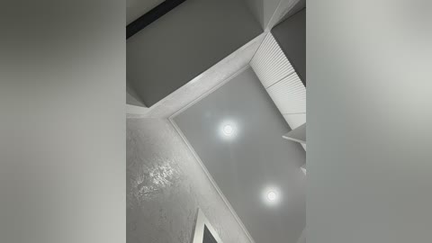 A black-and-white video of an angled ceiling with textured walls and three recessed lights, creating a modern, minimalist ambiance. The image emphasizes clean lines and geometric shapes.