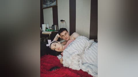 Video of a young couple in bed, the man shirtless and the woman in a pink headband, lying on a red, fluffy rug.