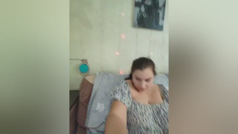 A blurry video of a woman with light skin and dark hair, wearing a striped top, sitting on a gray couch against a beige wall adorned with string lights and a black and white picture.