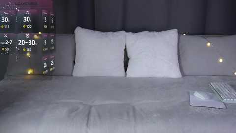 Video of a dimly lit bedroom with a grey bedspread, white pillows, and a white keyboard on the right. The background shows a dark, curtained window with a weather forecast overlay.