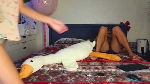 Video of a man and woman, nude, lying on a bed with a large white duck toy, in a bedroom with patterned bedding and decor.