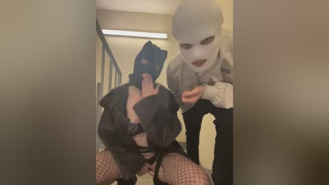 Video of a woman in a black leather jacket, fishnet stockings, and a black mask, being restrained by a man in a white balaclava and police uniform, in a dimly lit corridor.
