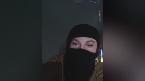 Video of a person in dim lighting, wearing a black beanie, mask, and olive-green jacket, with their eyes partially visible, standing in front of a dark, shadowy background.