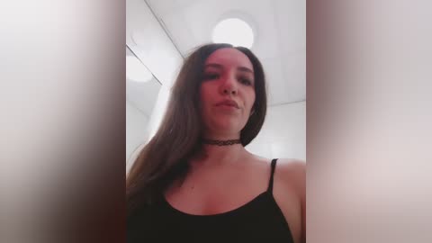 Video of a young woman with long brown hair, wearing a black spaghetti-strap top, standing in a white-walled room with circular ceiling lights. She has a black choker necklace.