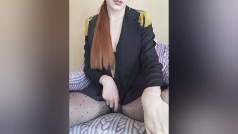 Video of a fair-skinned woman with long, straight auburn hair, wearing a black blazer with yellow epaulets, sitting on a patterned purple and white couch. She has sheer polka-dot stockings and a partially unbuttoned shirt.
