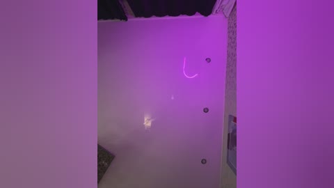 Media: A video of a dimly lit, unfinished interior room with white walls and a purple glow from a light source above, showing a bare concrete floor and exposed brick wall.