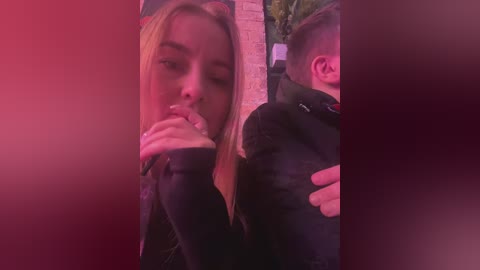 Media: Video of a blonde woman in a black jacket drinking from a pink cup, standing in a dimly lit room with a red tint.