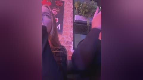 Media: Video of a young woman with long, straight hair wearing a black jacket, partially obscured by a brick wall and foliage, under dim, purple-hued lighting.