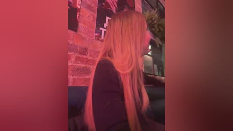 Media: Video of a blonde woman with long, straight hair, seated with her back to the camera in a dimly lit, brick-walled room, wearing a black jacket, with pink and purple lighting creating a soft, romantic ambiance.