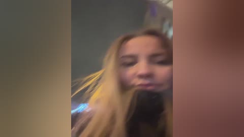 Media: A blurry video of a young woman with long blonde hair, wearing a dark jacket, standing indoors, partially obscured by vertical objects.