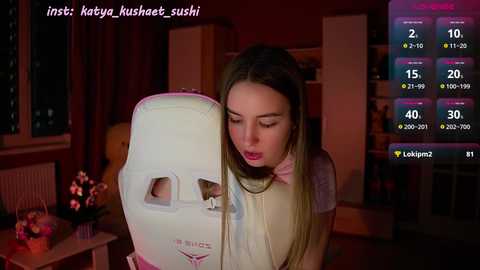 Media: Video of a young woman with long hair, wearing a beige sweater, leaning against a white baby stroller. The background shows a dimly lit living room with red walls, a window, and a pink flower arrangement.