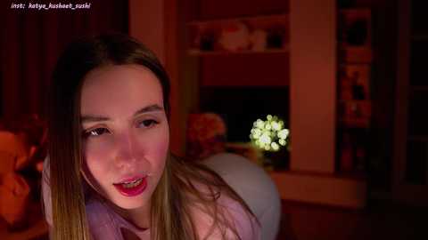 Media: Video of a smiling Asian woman with straight, light brown hair, wearing red lipstick and a light purple top, in a dimly lit room with a bookshelf and a small, glowing light in the background.