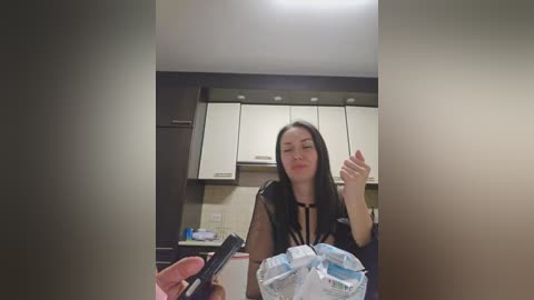 Video of a woman with long black hair and a tattoo on her arm, holding a white plastic bag with a blue water bottle inside. She stands in a modern kitchen with white cabinets and a black countertop.
