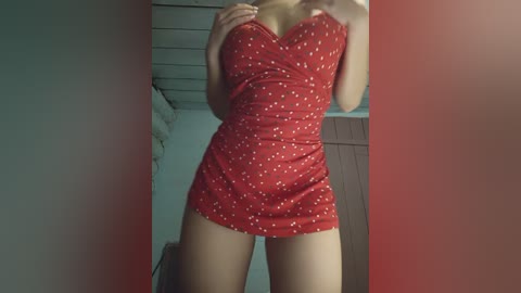 Video of a light-skinned woman in a tight, red mini-dress with white polka dots, standing in a dimly lit room with wooden paneling and a bed visible in the background.