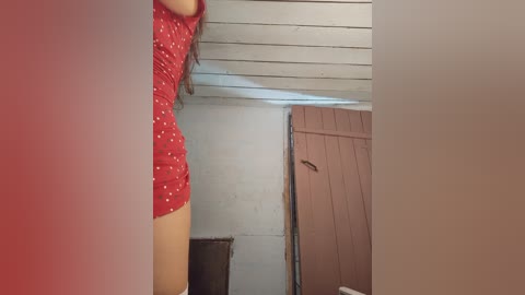 Video of a woman in a red, polka-dotted dress standing in a rustic, dimly lit room with white walls, wooden ceiling, and brown wooden door.