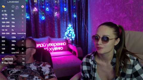 A video of a woman with light skin and brown hair, wearing sunglasses and a plaid shirt, sitting next to a man in a camouflage shirt, in a dimly-lit room with neon lights and a Christmas tree.