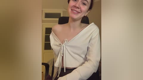Media: A video of a smiling young woman with short dark hair, wearing a white off-shoulder crop top and black pants, seated indoors.