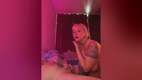 Video of a topless, fair-skinned, tattooed woman with short blonde hair, posing on a bed with a colorful blanket. Room has red and pink lighting, a dark curtain, and a mirror.