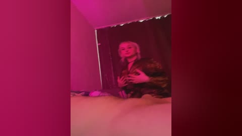 A video of a blonde woman in a dark room with pink lighting, wearing a dark robe, sitting on a bed with a white sheet.