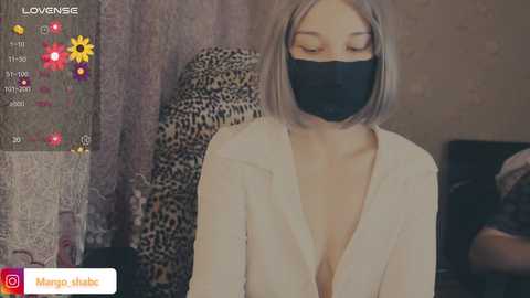 A video of a young woman with a short, platinum blonde bob, wearing a black face mask and a white, low-cut blouse, sitting in a leopard-print chair. The background features a textured wall and a colorful social media overlay.