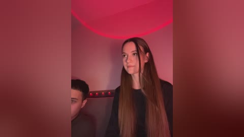Media: Video of a woman with long brown hair, wearing a black top, standing in a dimly lit room with red lighting. The background is abstract with blurred shapes.