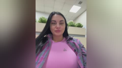 A video of a young woman with long black hair, wearing a pink top and a plaid jacket, smiling in an office setting with green plants in the background.