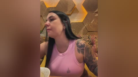 A video shows a tattooed woman in a light pink tank top, eating at a restaurant with a geometric, golden-patterned wall.