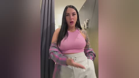 Video of a young woman with long black hair, wearing a pink crop top and gray high-waisted pants, standing in a dressing room with a gray curtain.