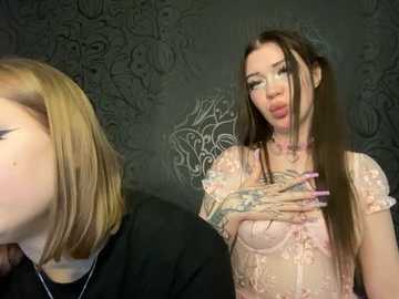 Media: Video of a pale-skinned woman with long, dark hair, wearing a sheer, pink lace top, standing against a dark, ornate wallpaper. She covers her chest with one hand. Another woman with shoulder-length, light hair is partially visible in the foreground.