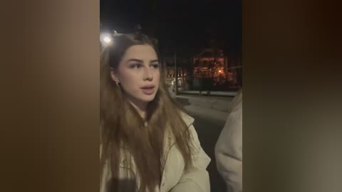 Media: Video of a young woman with long, wavy brown hair, wearing a beige jacket, walking at night on a city street. Background includes dimly lit buildings and streetlights.