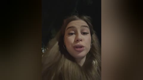 Media: Video of a young woman with light skin, straight brown hair, and a fur-lined coat, puckering her lips, taken indoors with dim lighting.