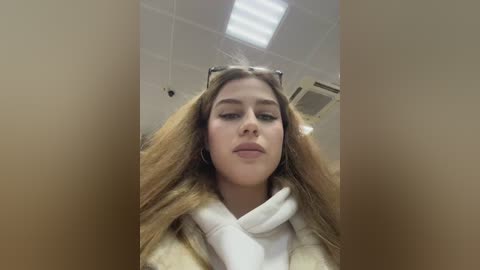 Media: Video of a young Caucasian woman with long, light brown hair, wearing a white coat and gloves, standing indoors under fluorescent lighting. She has a neutral expression and is partially obscured by the camera's focus.