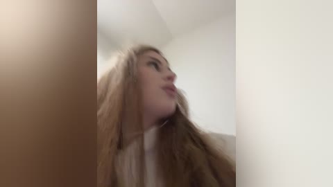 Media: A blurry video of a woman with long, light brown hair and pale skin, wearing a white top, standing indoors against a plain, white wall.