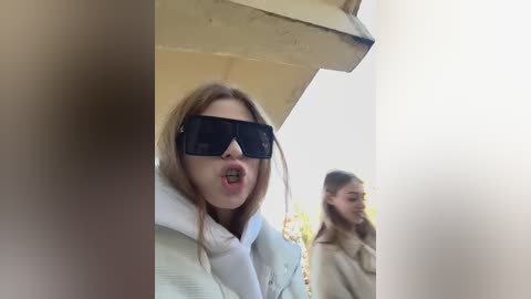 Media: Video of a blonde woman wearing large black sunglasses, open-mouthed, against a blurred background, possibly a beach scene with another person partially visible.