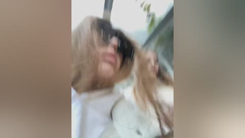 Media: A blurry video of a blonde woman with sunglasses and a white shirt, captured in a car with a blurred background, likely driving.