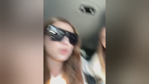 Media: Video of a woman with long, straight brown hair, wearing large, dark sunglasses and a white shirt, in a blurred, indoor setting with a ceiling light visible.