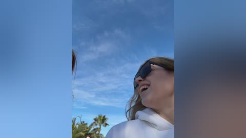 Media: Video of a blonde woman in sunglasses, laughing, with a feather in her mouth, against a bright blue sky with wispy clouds and palm trees.