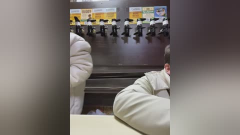 Media: Video of a barbershop with a row of black barber chairs and a row of yellow and white \"Dove\" shampoo bottles overhead. A customer with short brown hair sits in the foreground, wearing a beige jacket.