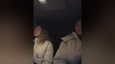 Media: Video of a dimly-lit room showing two people, a woman with curly hair and a man with a bald head, both wearing beige coats, facing away from the camera.